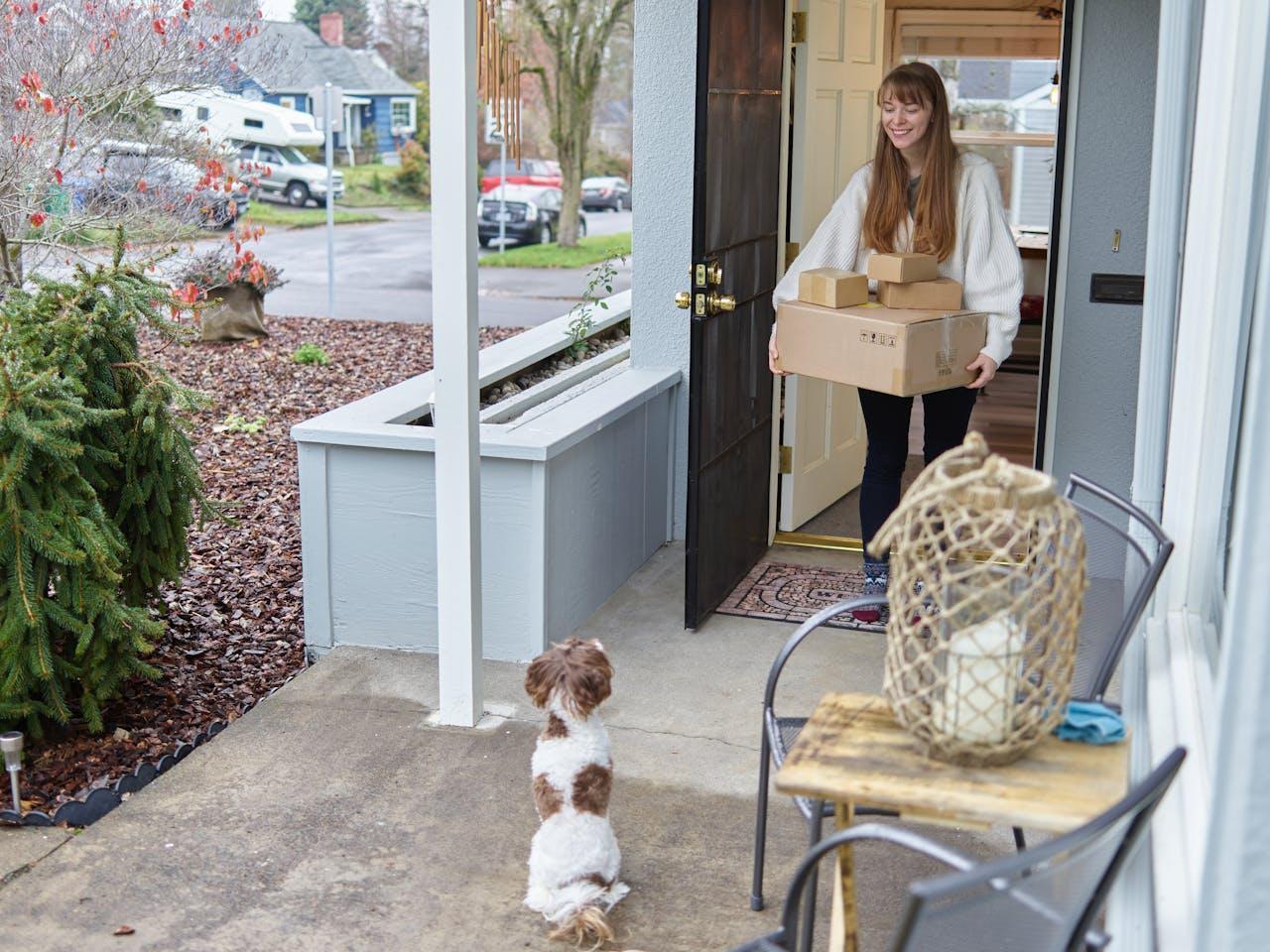A woman standing in the doorway holding boxes near a white and brown dog,